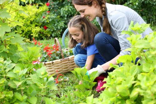 A woman and a young girl are gardening together in a lush, well-maintained backyard, surrounded by dense green foliage and various flowering plants. The woman, with long braided hair, is kneeling on the grass, leaning forward while assisting the girl, who is wearing a blue top and jeans. They are both carefully tending to a flower bed, which includes a mix of flowering shrubs with red blossoms and leafy green plants. In the foreground, there is a small section of neatly trimmed grass with a smooth, dark soil border lining the flower beds. The background features a combination of taller bushes and trees, providing a shaded and natural environment. A wicker basket rests nearby, possibly for collecting garden produce or weeds. The scene appears to be a warm, sunny day, ideal for outdoor gardening activities. This image exemplifies the type of outdoor garden maintenance and plant care services offered by Gardening Services Woolwich in the Woolwich area, highlighting professional attention to garden design and upkeep.